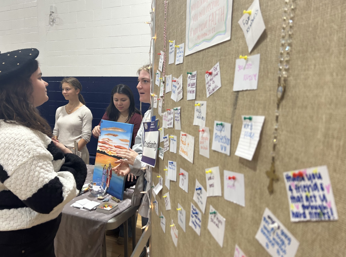 Senior Finn Skobel (right) talks with a viewer of the Christianity booth. To the right of her is the prayer board –the notes on which are filled with prayer requests from fellow students– many reading “strength” and “protection.”